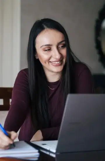 Smiling woman writing notes while using a laptop, representing virtual interviews hiring.