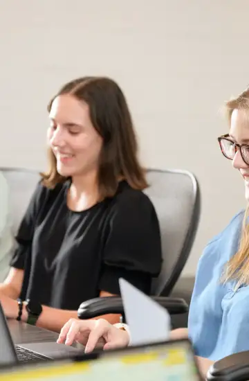 Three people are seated at a table working on laptops; they are smiling and appear to be in a collaborative setting.