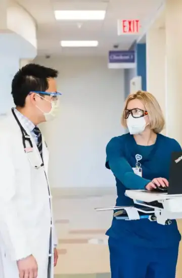 Two healthcare professionals wearing masks and scrubs discussing data on a hospital laptop cart.