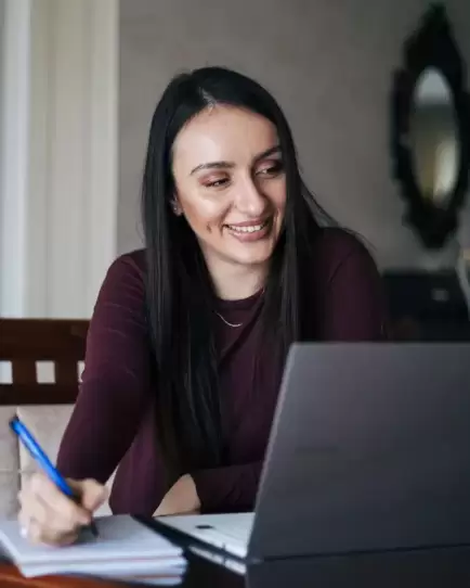 Smiling woman writing notes while using a laptop, representing virtual interviews hiring.
