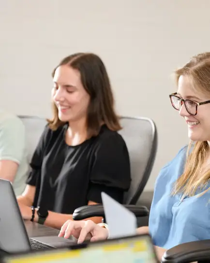 Three people are seated at a table working on laptops; they are smiling and appear to be in a collaborative setting.