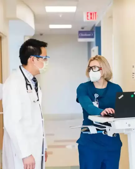 Two healthcare professionals wearing masks and scrubs discussing data on a hospital laptop cart.