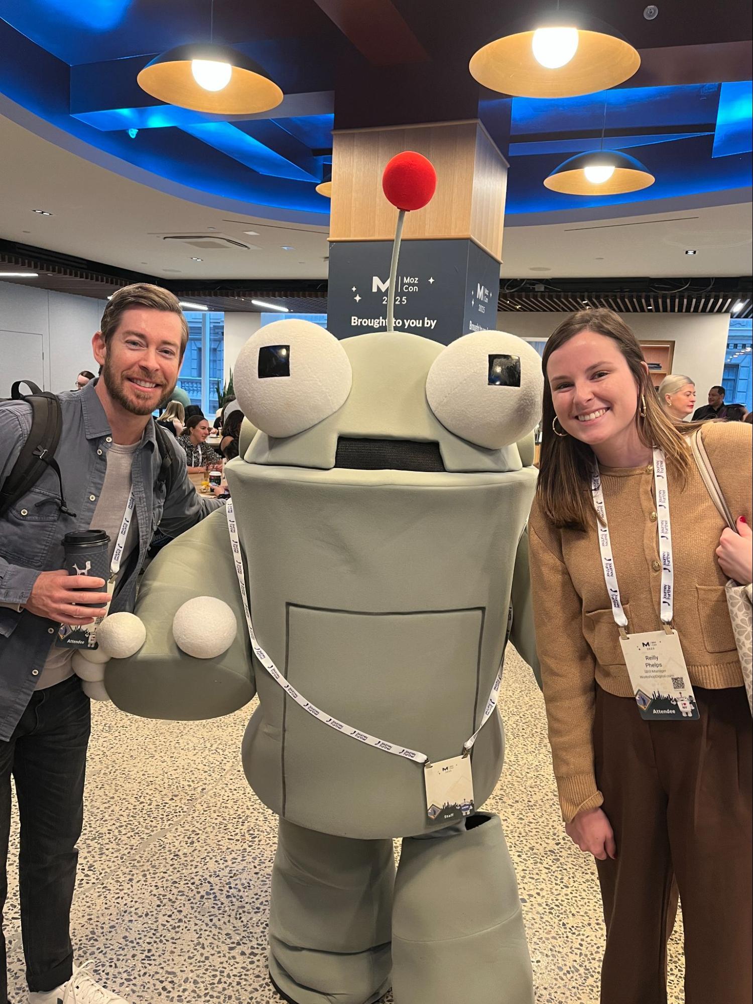 Brian and Reilly smiling and posing with the large green MozBot mascot at MozCon, standing in a busy conference hallway.